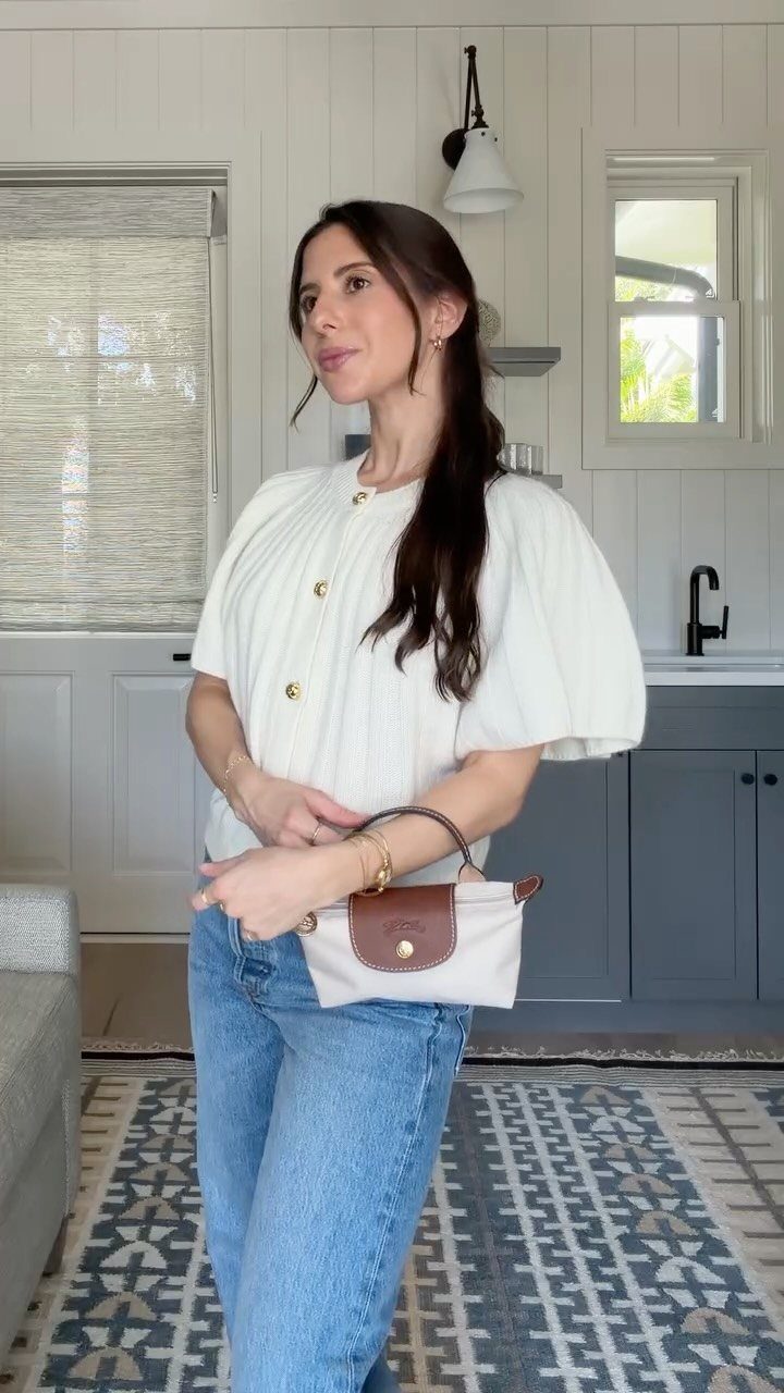Woman in a white buttoned blouse and blue jeans holding a small handbag.
