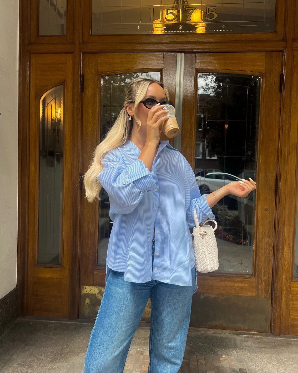 Blonde woman in a striped blue shirt and jeans holding a coffee cup outdoors.