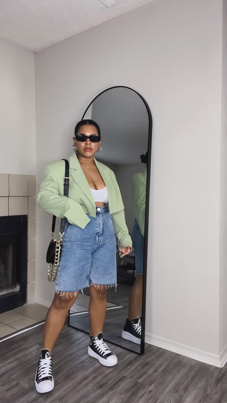 Woman posing in denim Bermuda shorts, a green blazer, and high-top sneakers.