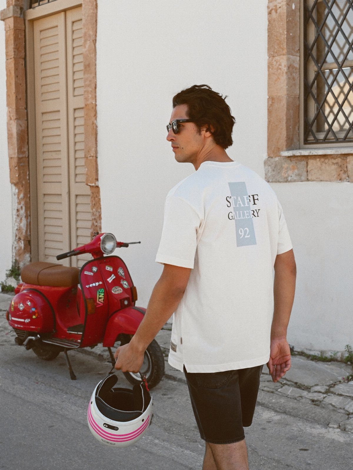 Man in a white graphic t-shirt and black shorts walking past a red scooter.