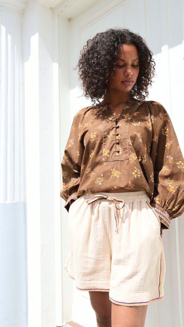 Woman wearing a brown floral blouse and cream linen drawstring shorts standing outdoors.
