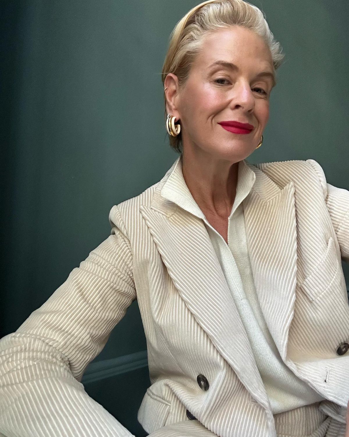 Blonde woman in a cream corduroy blazer and white collared sweater posing against a teal wall.
