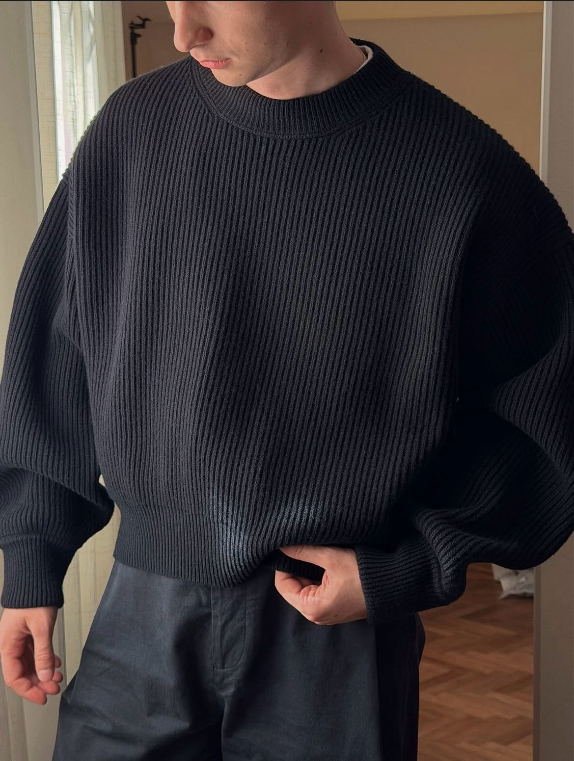 A person wearing a black ribbed oversized crewneck sweater and dark trousers indoors.