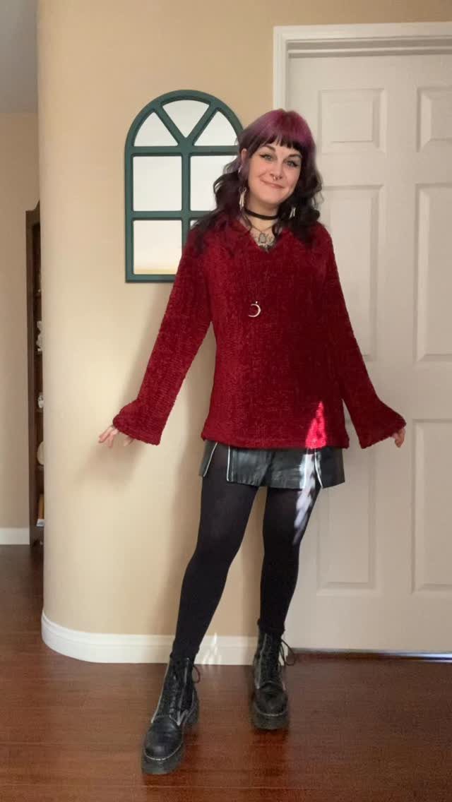 Woman posing in a red sweater, black leather skirt, black tights, and combat boots.