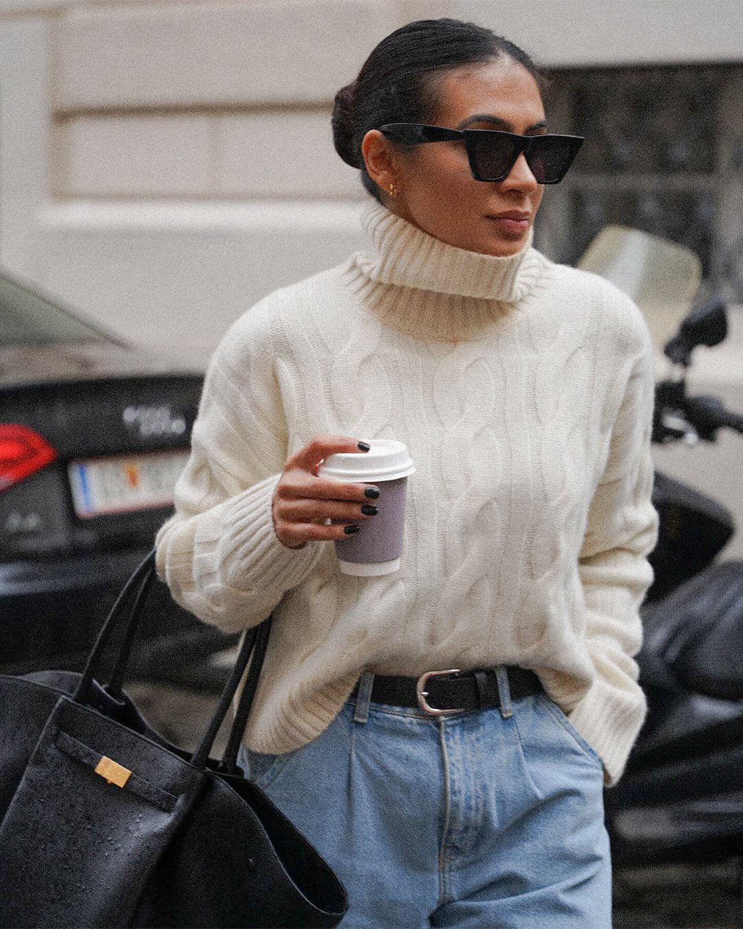 A woman in a cream cable-knit turtleneck sweater and blue jeans holding a coffee cup.