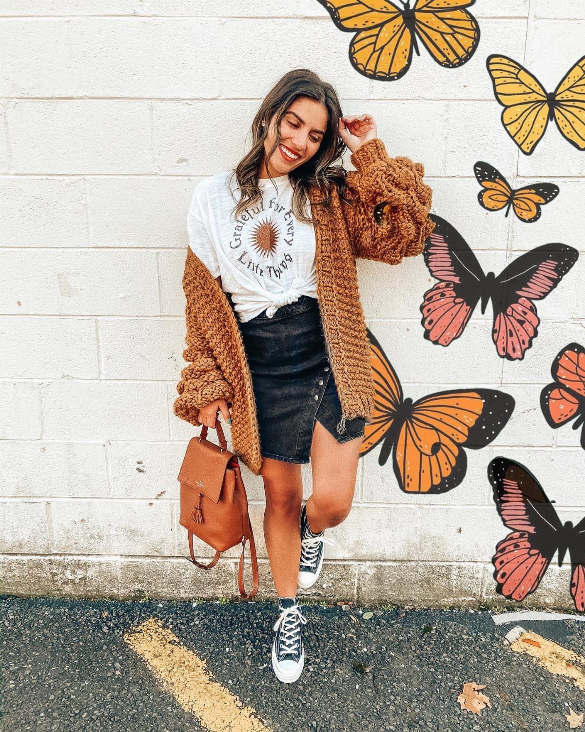 Smiling woman wearing a white graphic t-shirt, brown cardigan, and black denim mini skirt against a butterfly mural.