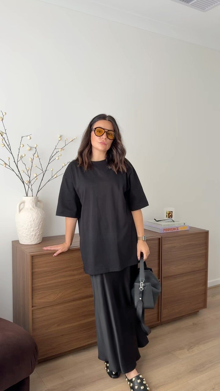 Woman posing in an oversized black t-shirt and a long black satin slip skirt.