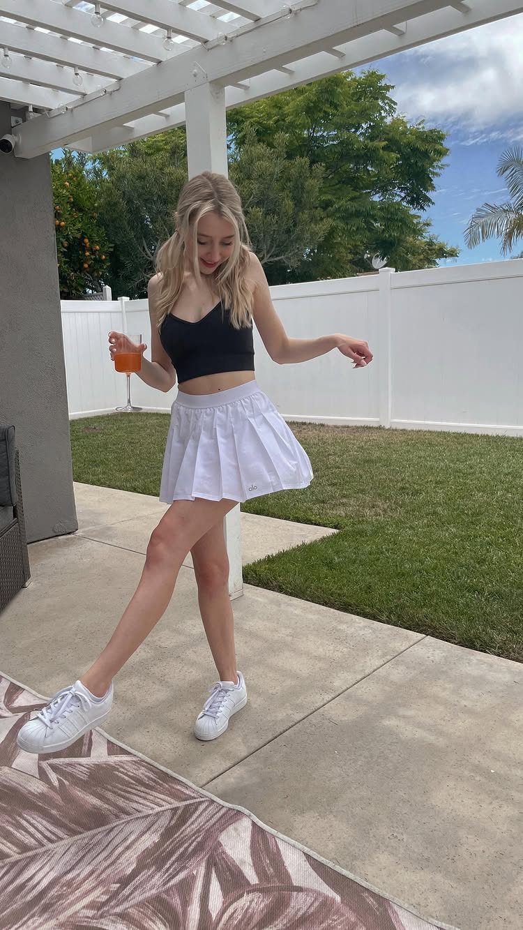 Blonde woman wearing a black crop top, white pleated skirt, and white sneakers.