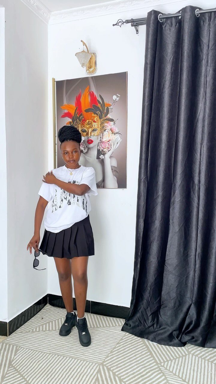 A young woman posing in a white graphic t-shirt, black pleated skirt, and black sneakers.