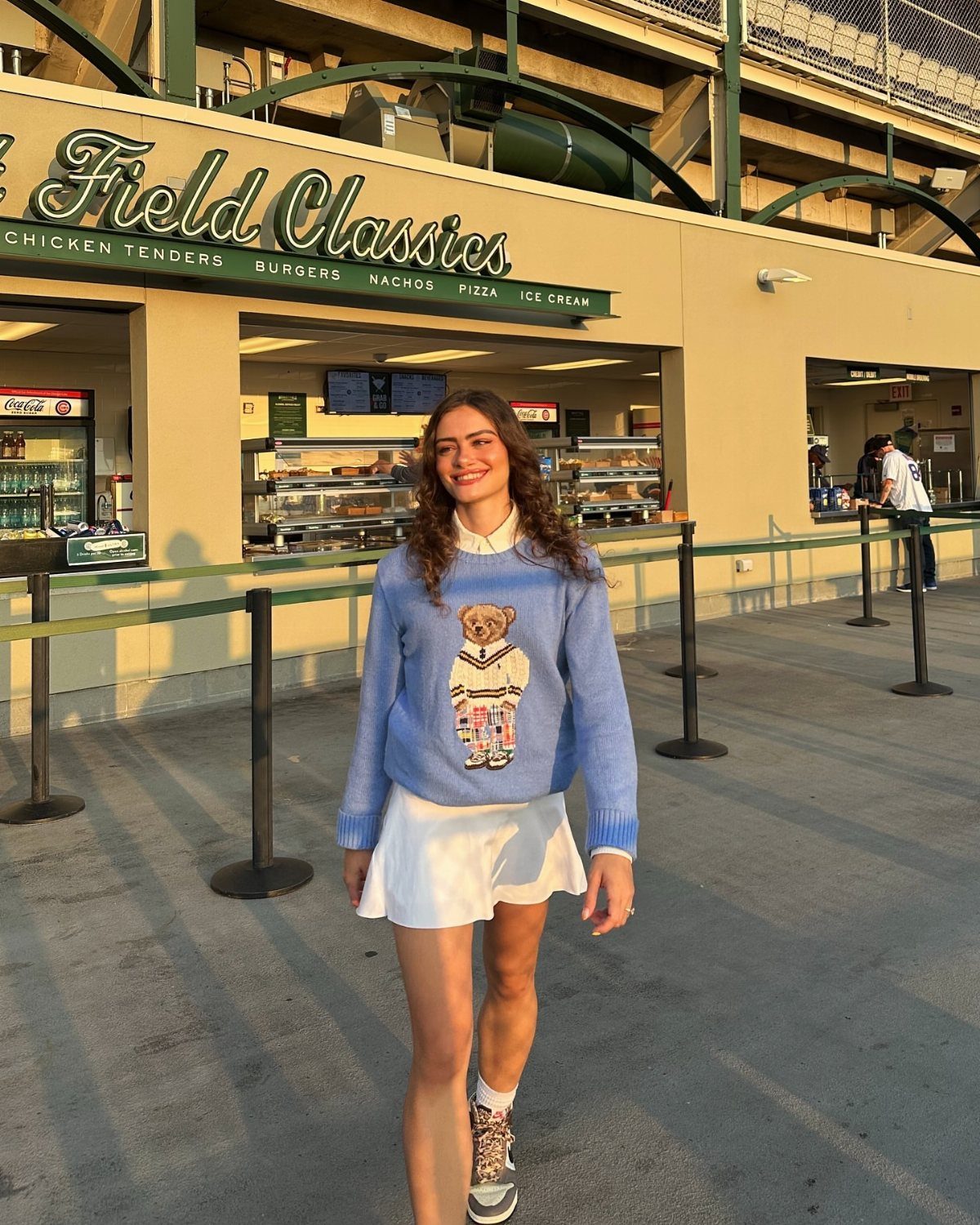 A smiling woman wearing a blue teddy bear sweater and a white tennis skirt walking outdoors.