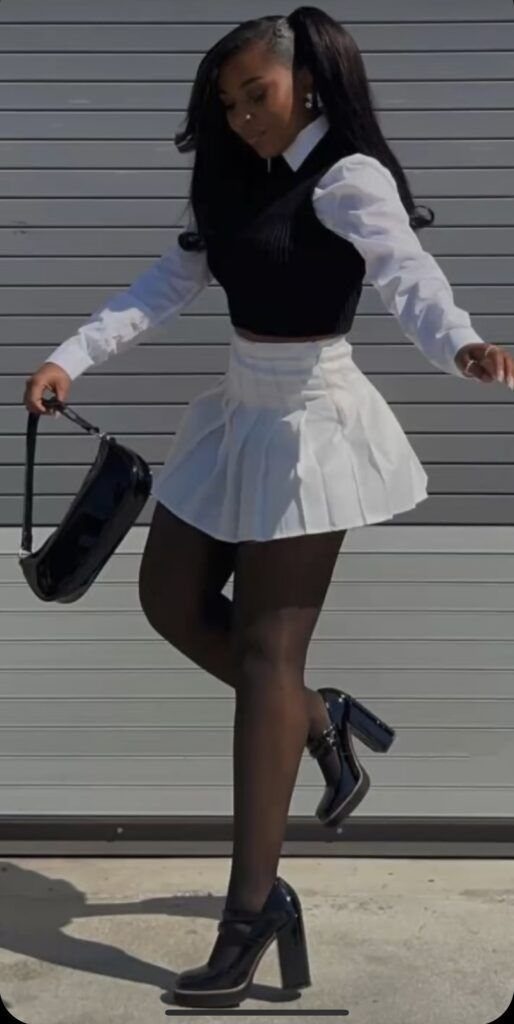 A woman in a white blouse, black vest, white pleated skirt, and black block heels.