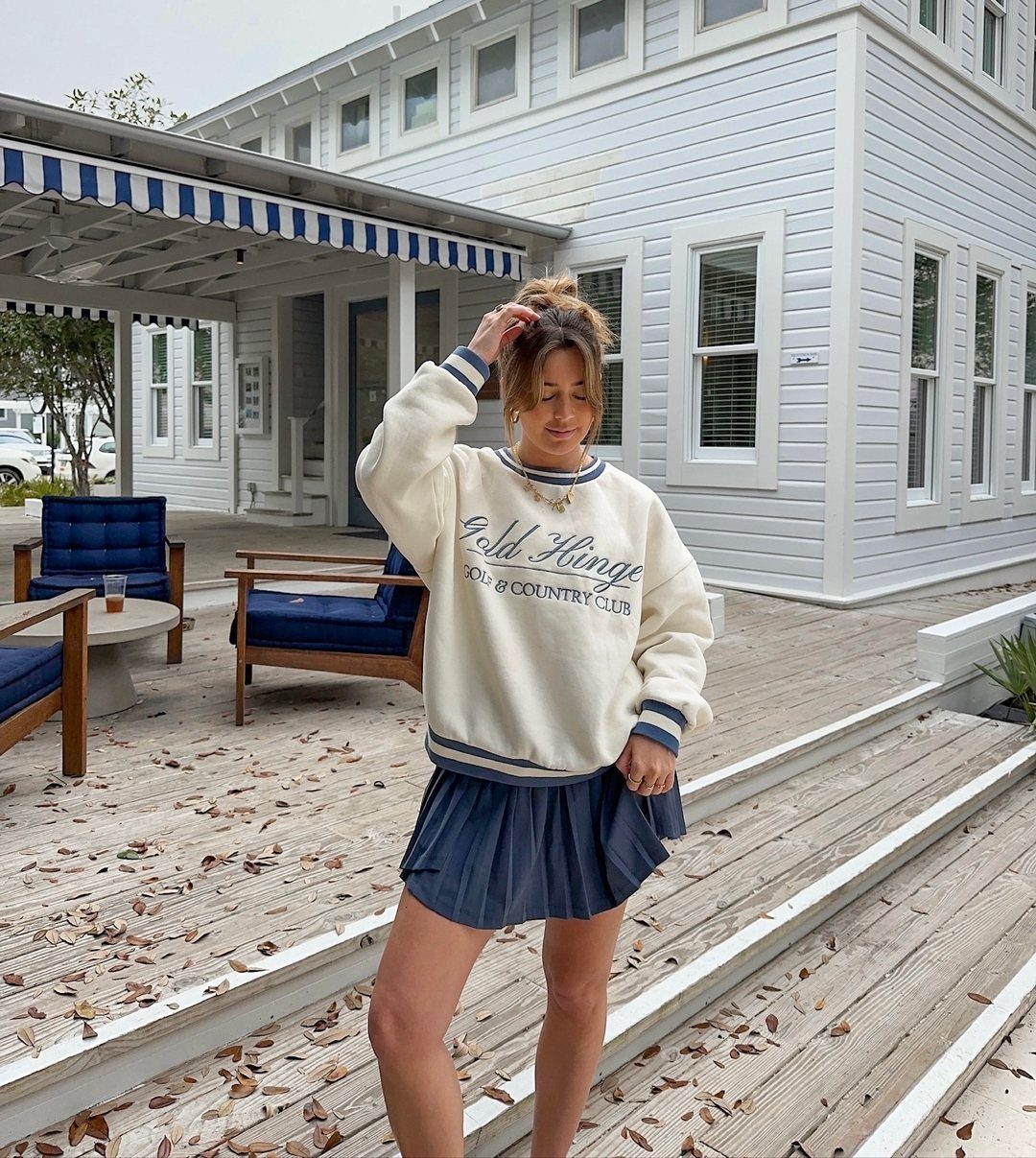 Woman wearing a cream sweatshirt and navy pleated tennis skirt standing on a wooden deck.
