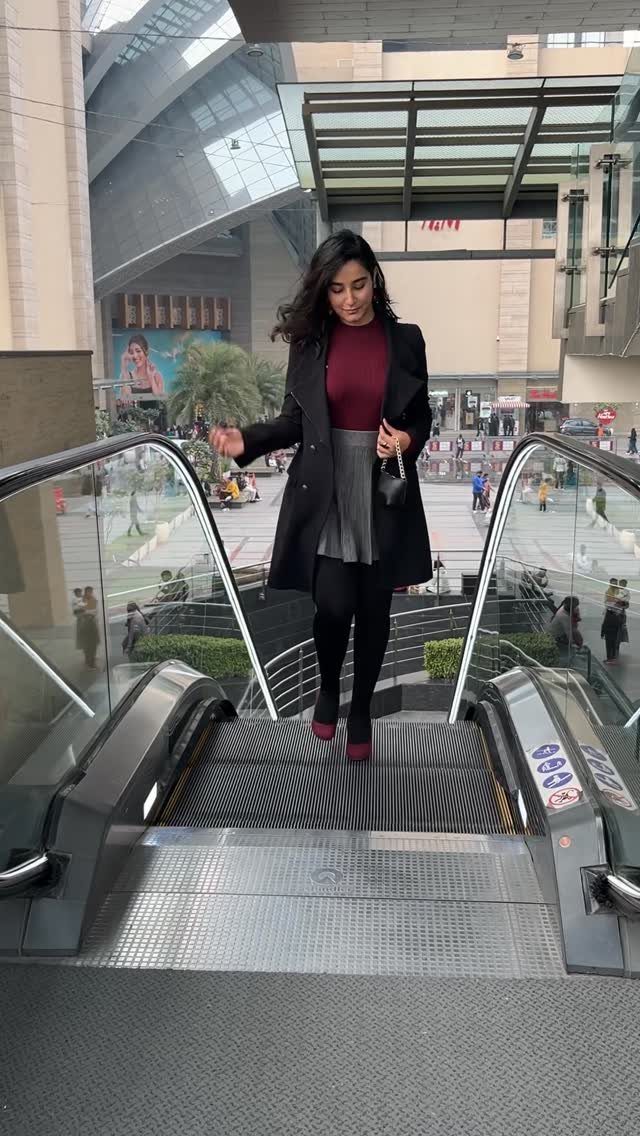 Woman wearing a dark turtleneck, grey tennis skirt, and long black coat on an escalator.
