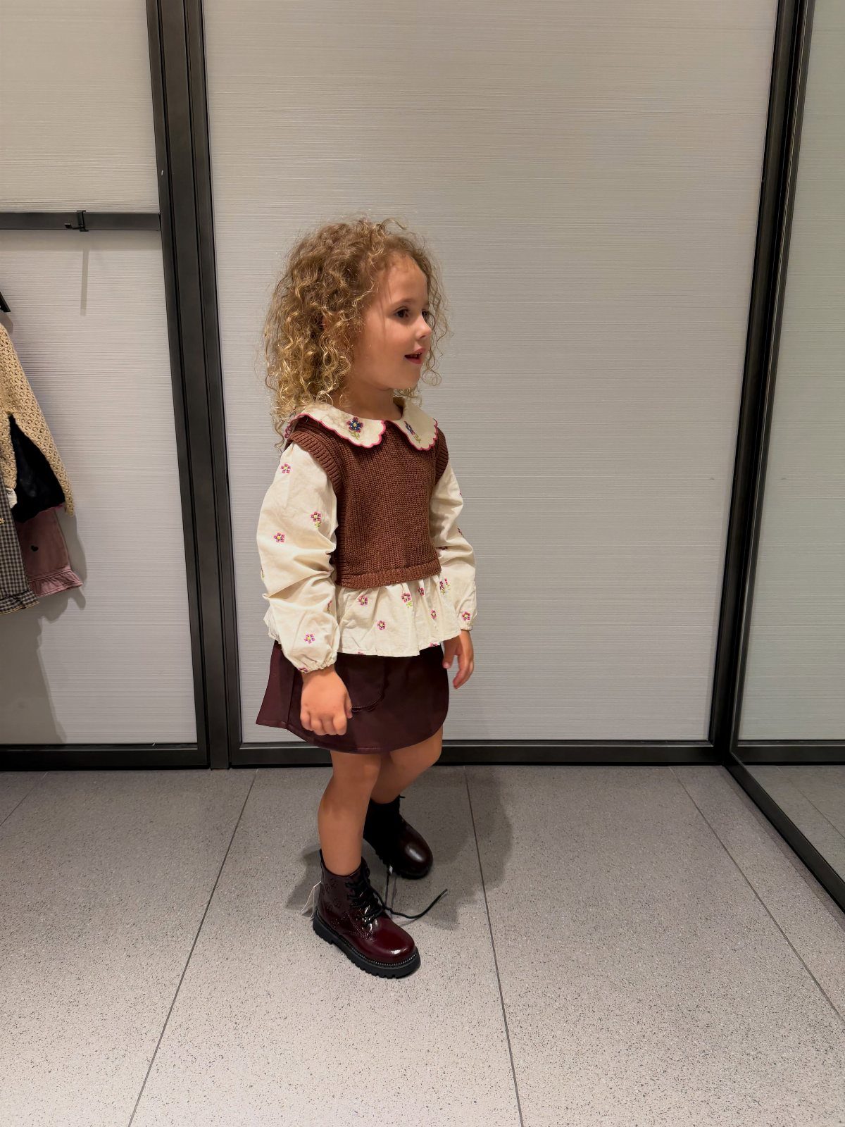Toddler girl with curly hair wearing a white blouse, brown vest, and dark skirt.