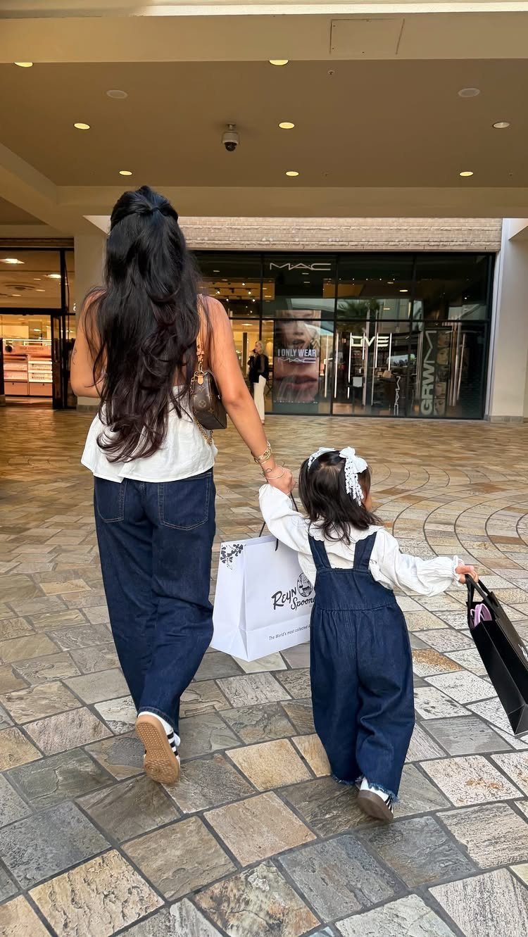 Toddler girl in denim overalls and white shirt walking through a shopping mall holding a bag.