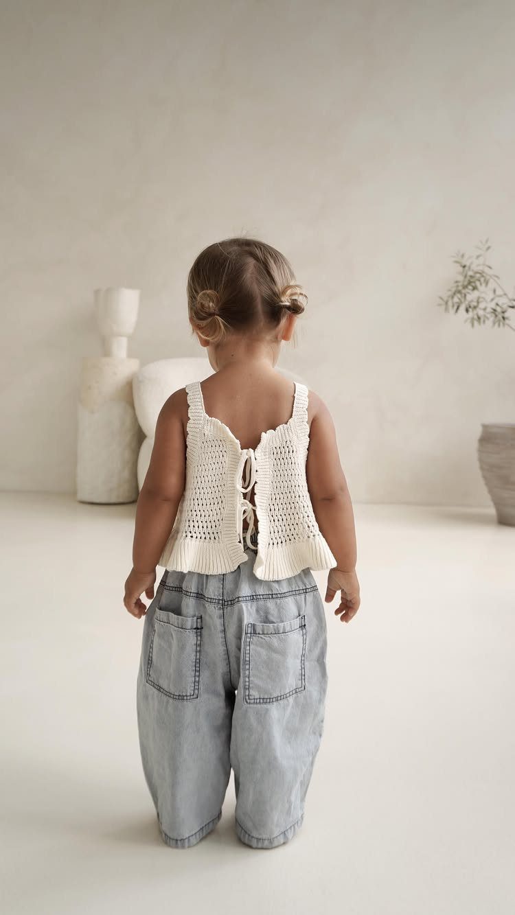 Toddler girl wearing a cream crochet vest and light blue denim trousers standing in a minimalist room.