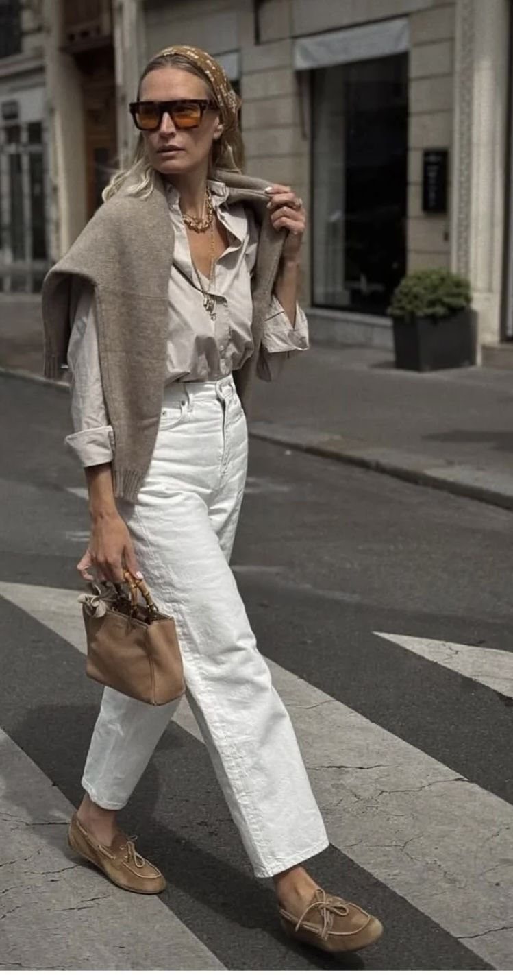 A woman walking in a street wearing white trousers, a beige shirt, and tan boat shoes.
