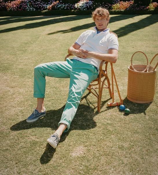 Man in white polo and teal trousers sitting on a lawn wearing boat shoes.