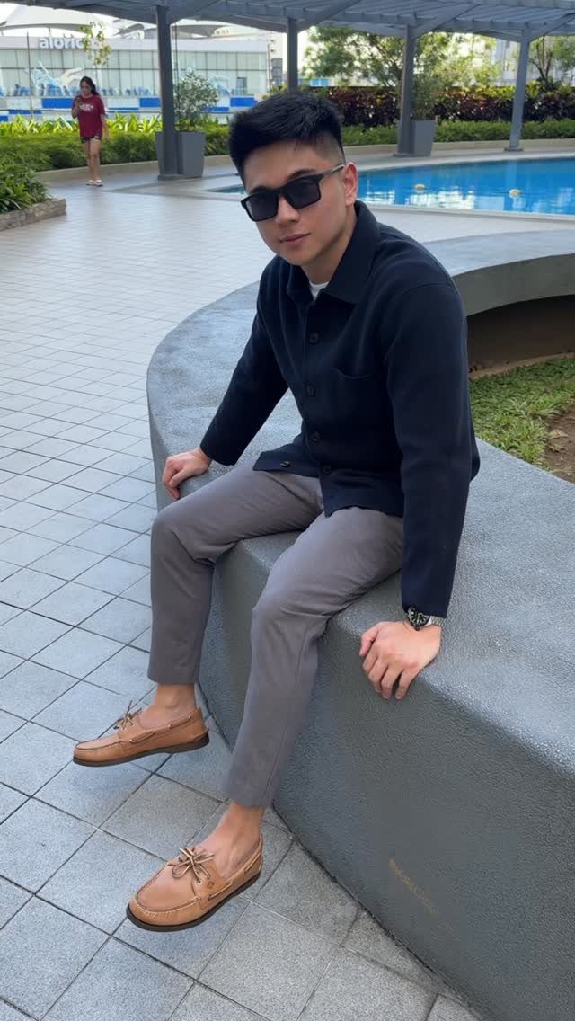 A man in a dark shirt and grey trousers wearing tan boat shoes sitting outdoors.