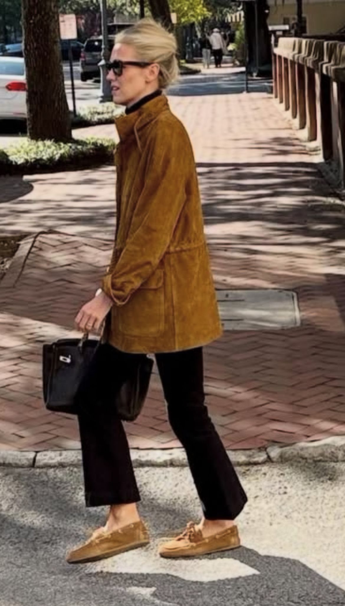 A woman walking in a brown suede jacket, black pants, and tan boat shoes.