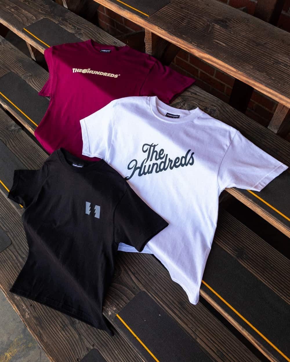 Three folded t-shirts in maroon, white, and black resting on dark wooden bleachers.
