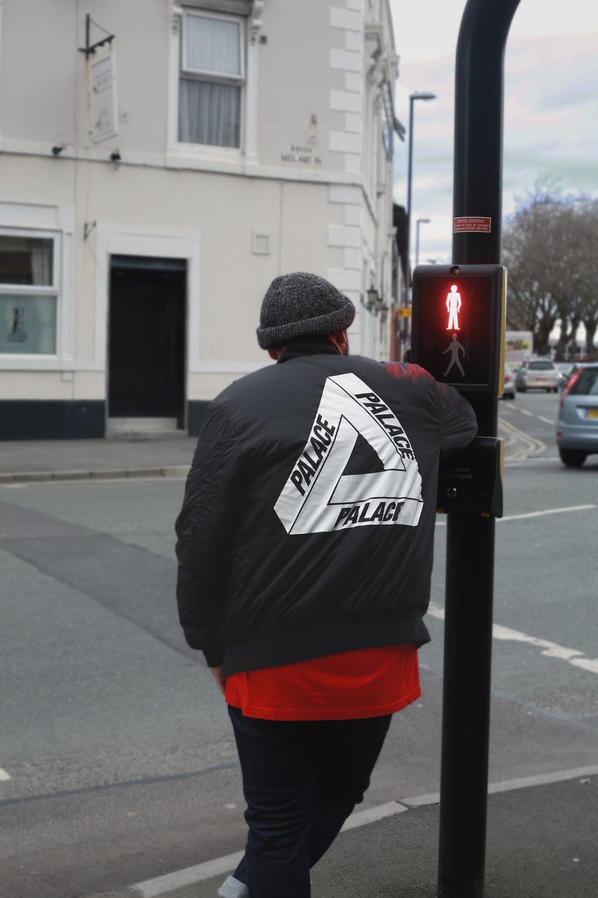 Person wearing a black Palace bomber jacket standing by a traffic light on a city street.