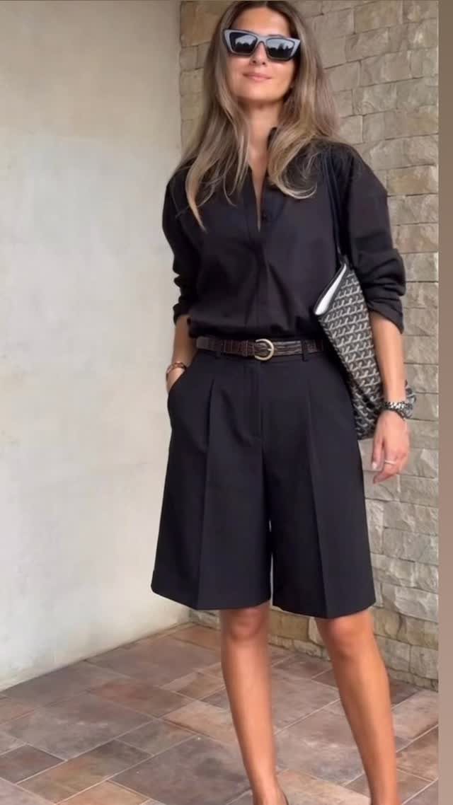 Woman wearing a black button-down shirt and tailored black Bermuda shorts with a brown belt.