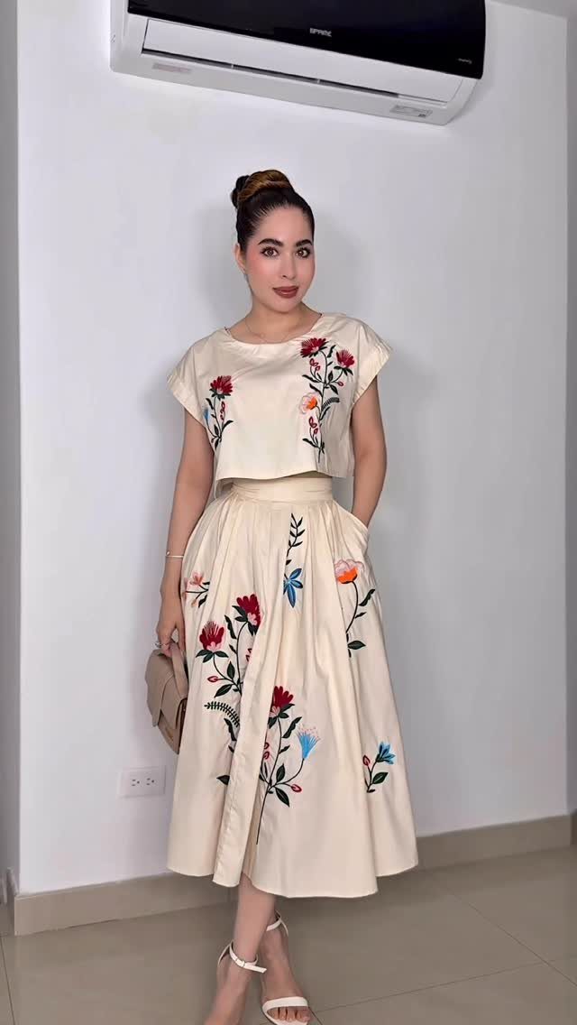 Woman in a cream two-piece outfit with colorful floral embroidery on the top and skirt.
