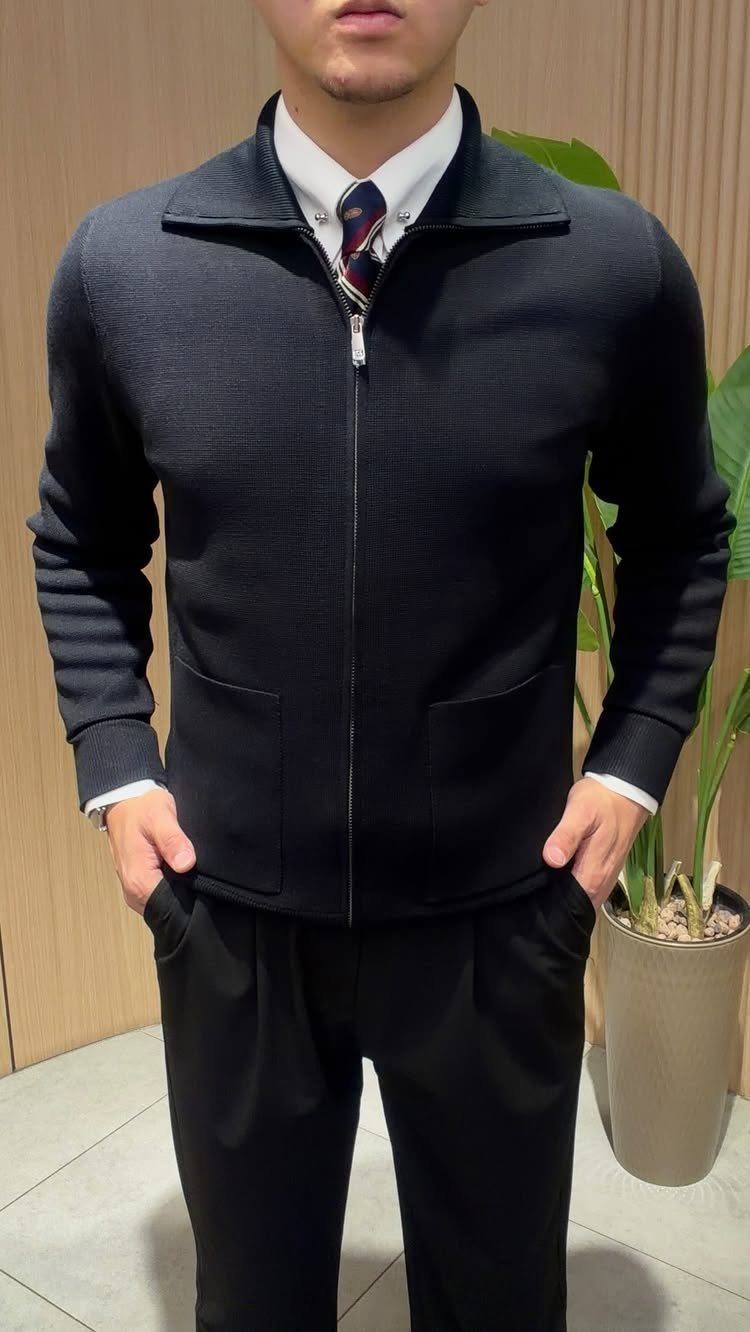 Man wearing a navy textured cardigan, white shirt, patterned tie, and black trousers.