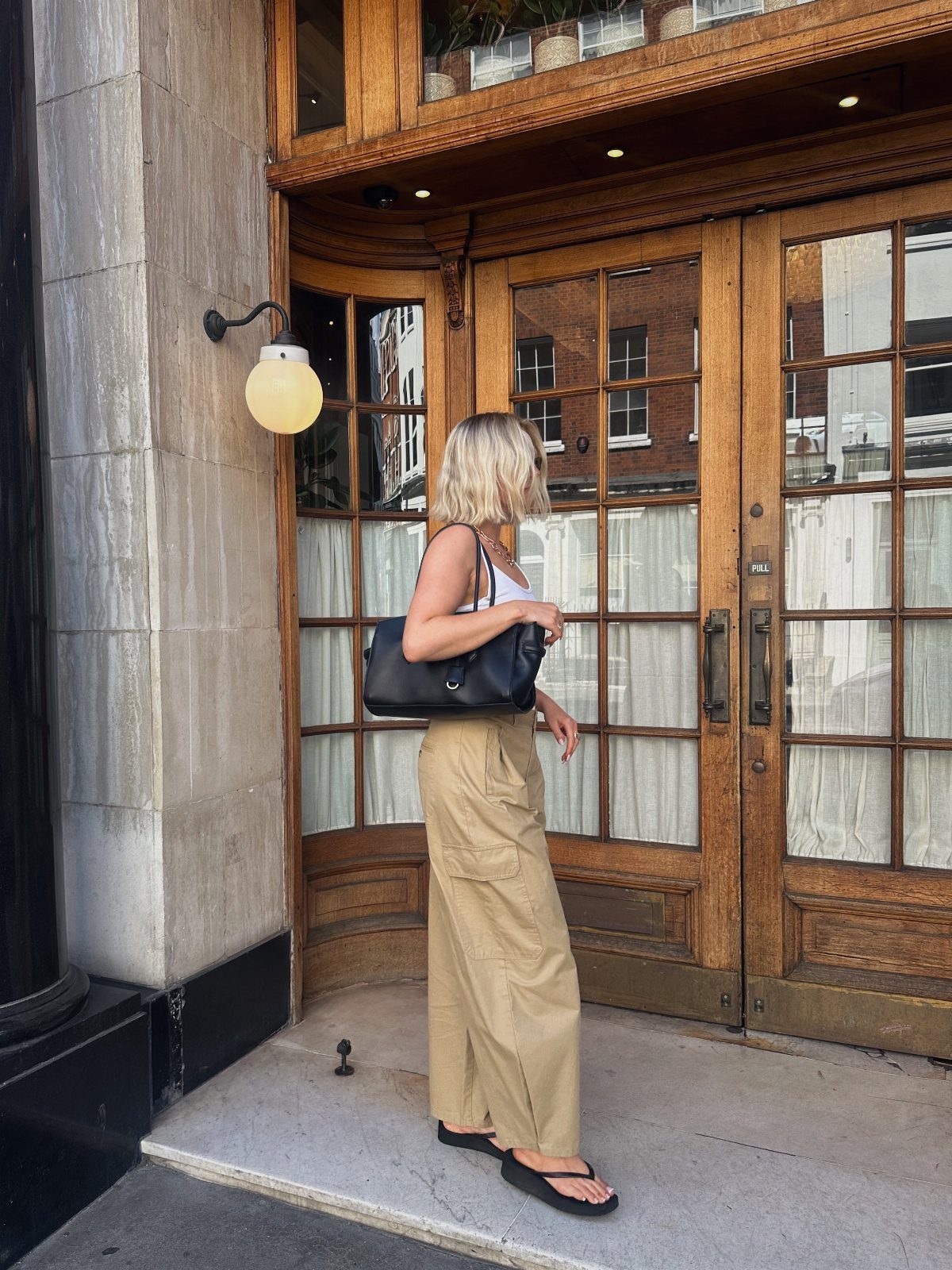 Blonde woman in white tank top and tan wide-leg trousers standing by wooden doors.