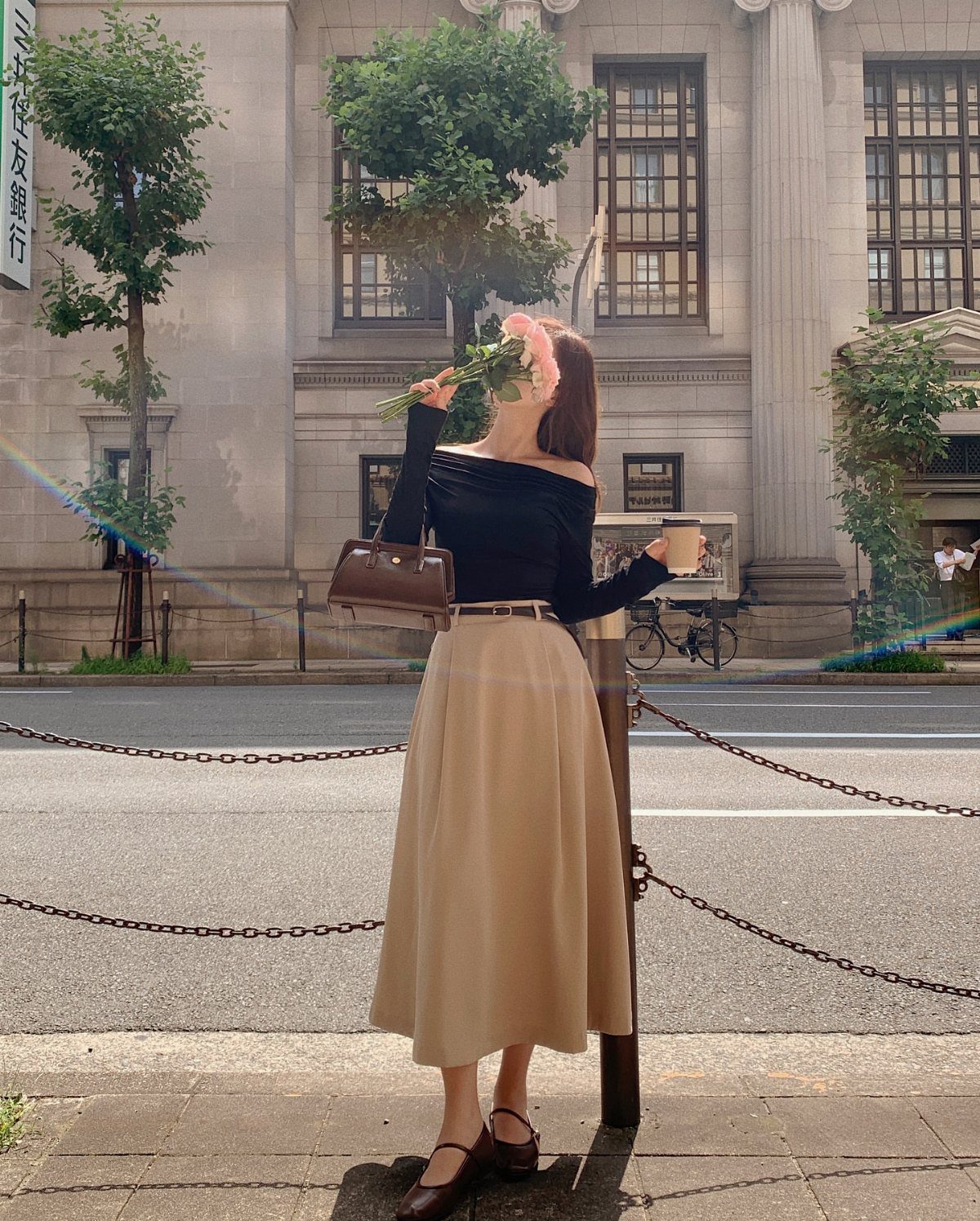 Woman in a black top and long beige skirt standing on a city street holding flowers and a coffee cup.
