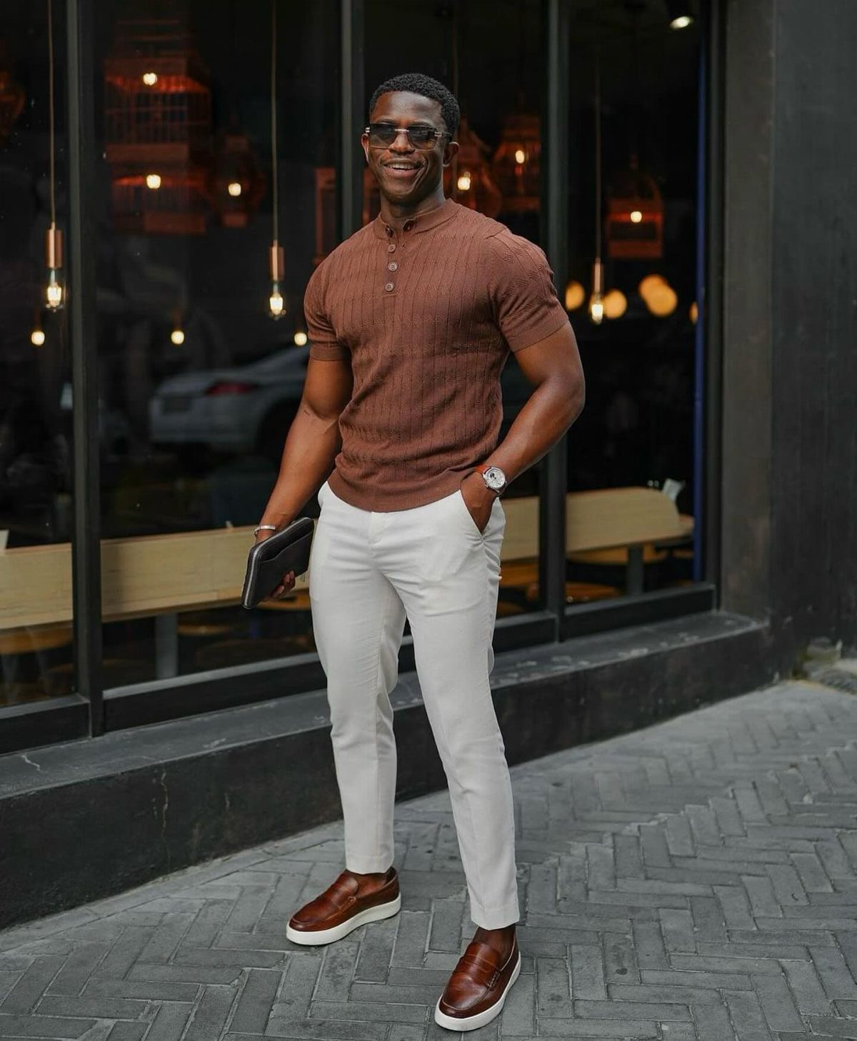 A man in a brown henley and white tailored trousers standing on a city sidewalk.