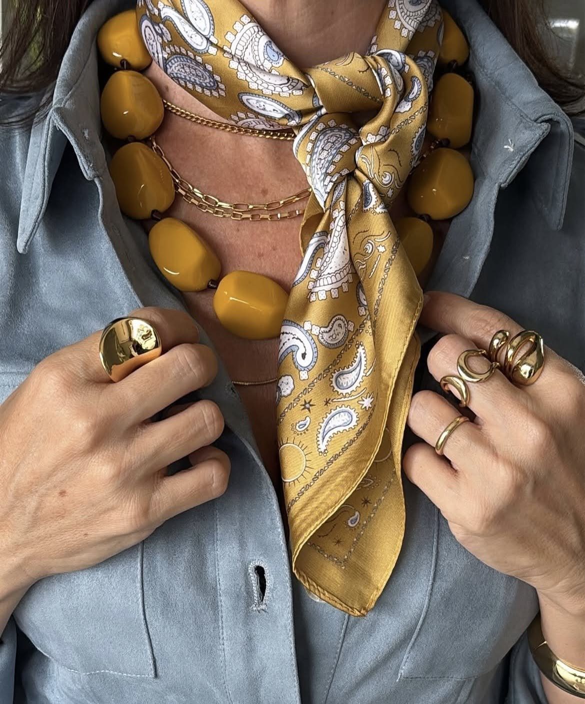 Person wearing a blue shirt with a yellow silk scarf, chunky bead necklace, and gold rings.