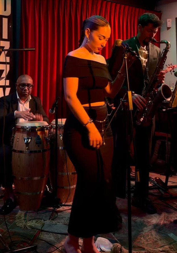 A woman in black attire stands in a jazz club with musicians performing in the background.