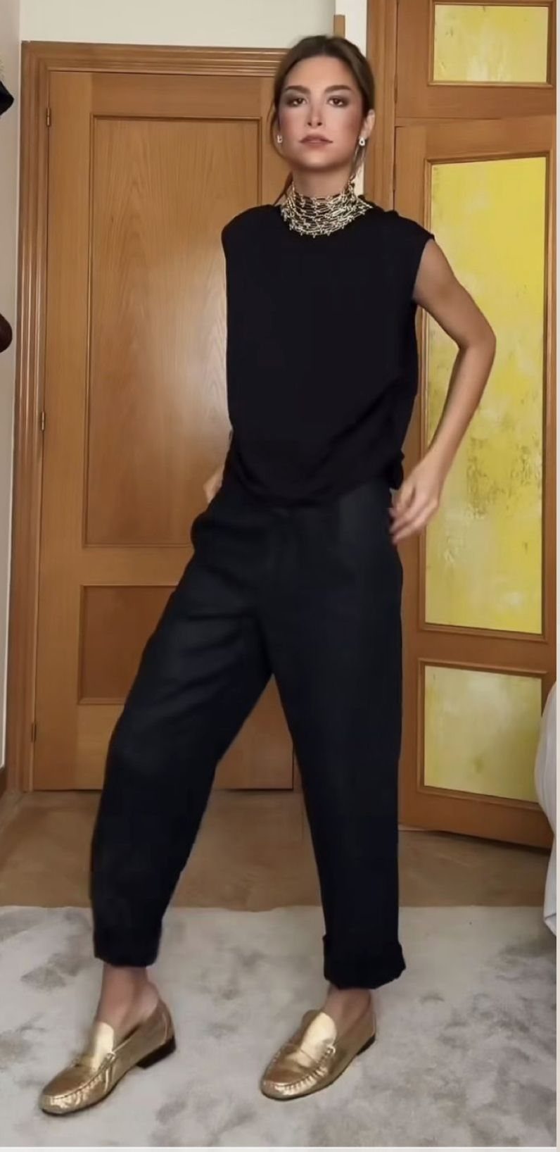 Woman posing in black sleeveless top, black trousers, and metallic gold loafers on a rug.
