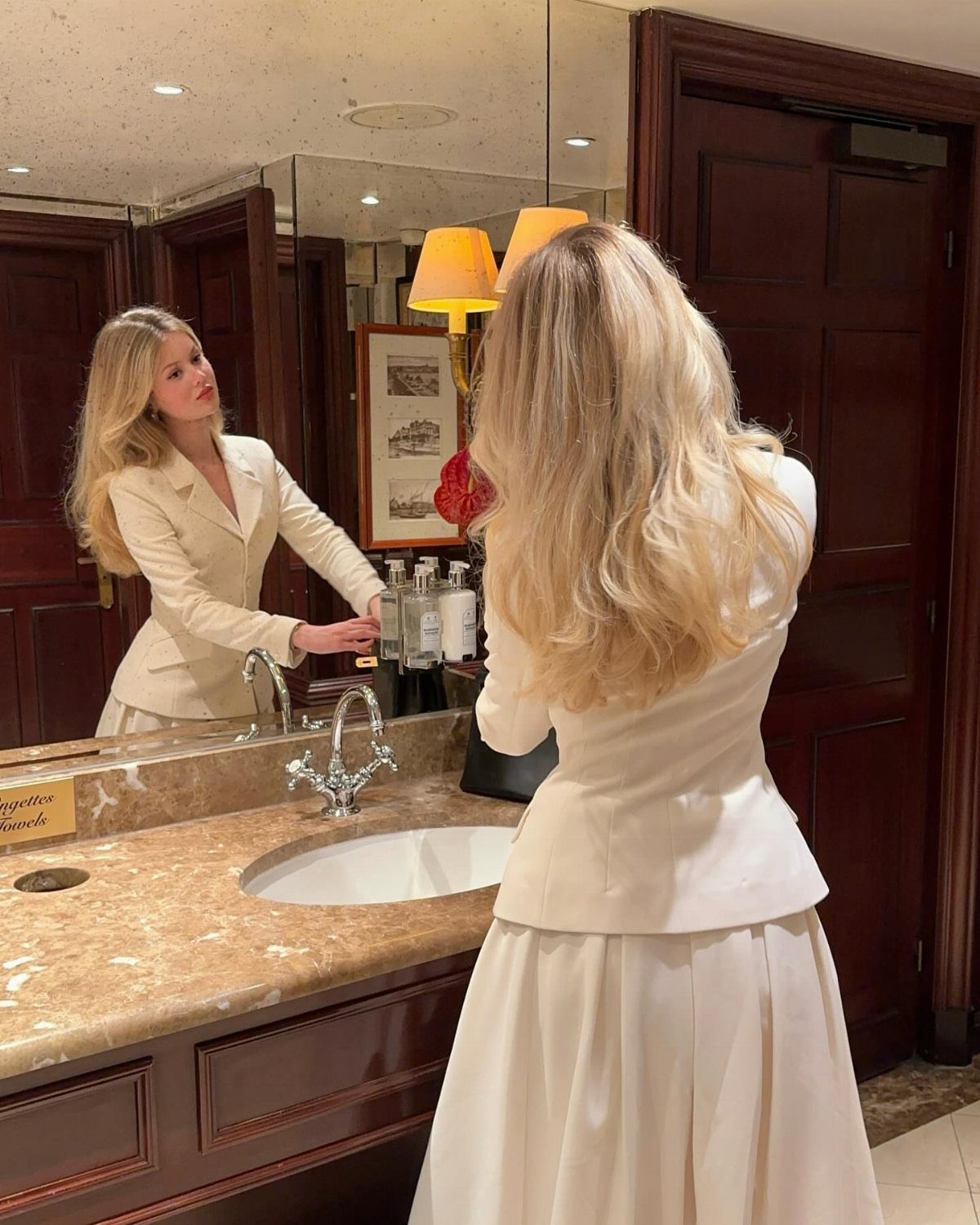 Blonde woman in a cream tailored blazer and skirt looking into a bathroom mirror.
