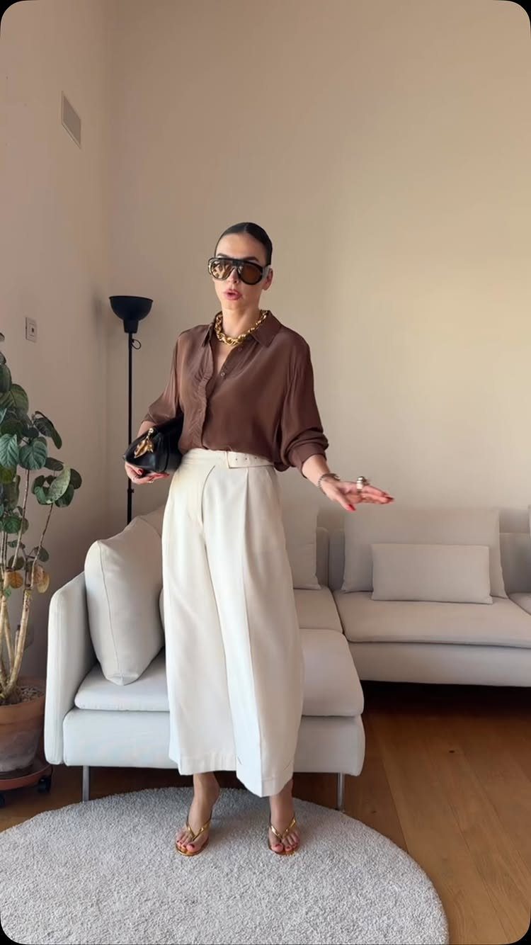 Woman in brown shirt and cream trousers accessorized with gold jewelry and sunglasses.