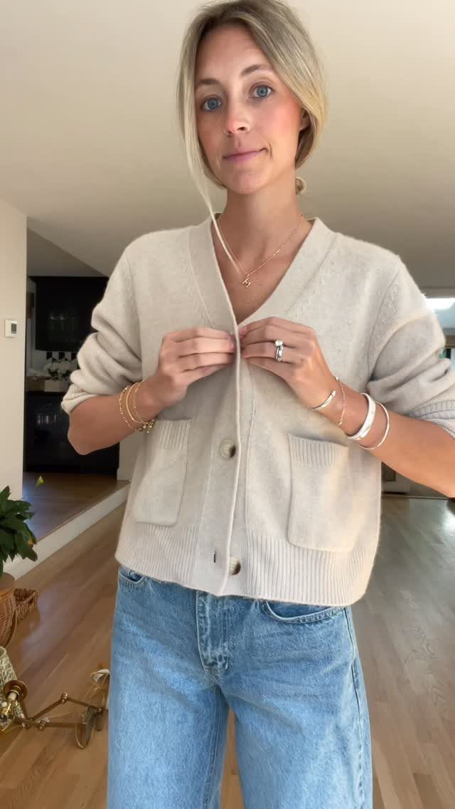 Blonde woman wearing a cream knit cardigan and blue jeans standing in a bright room.