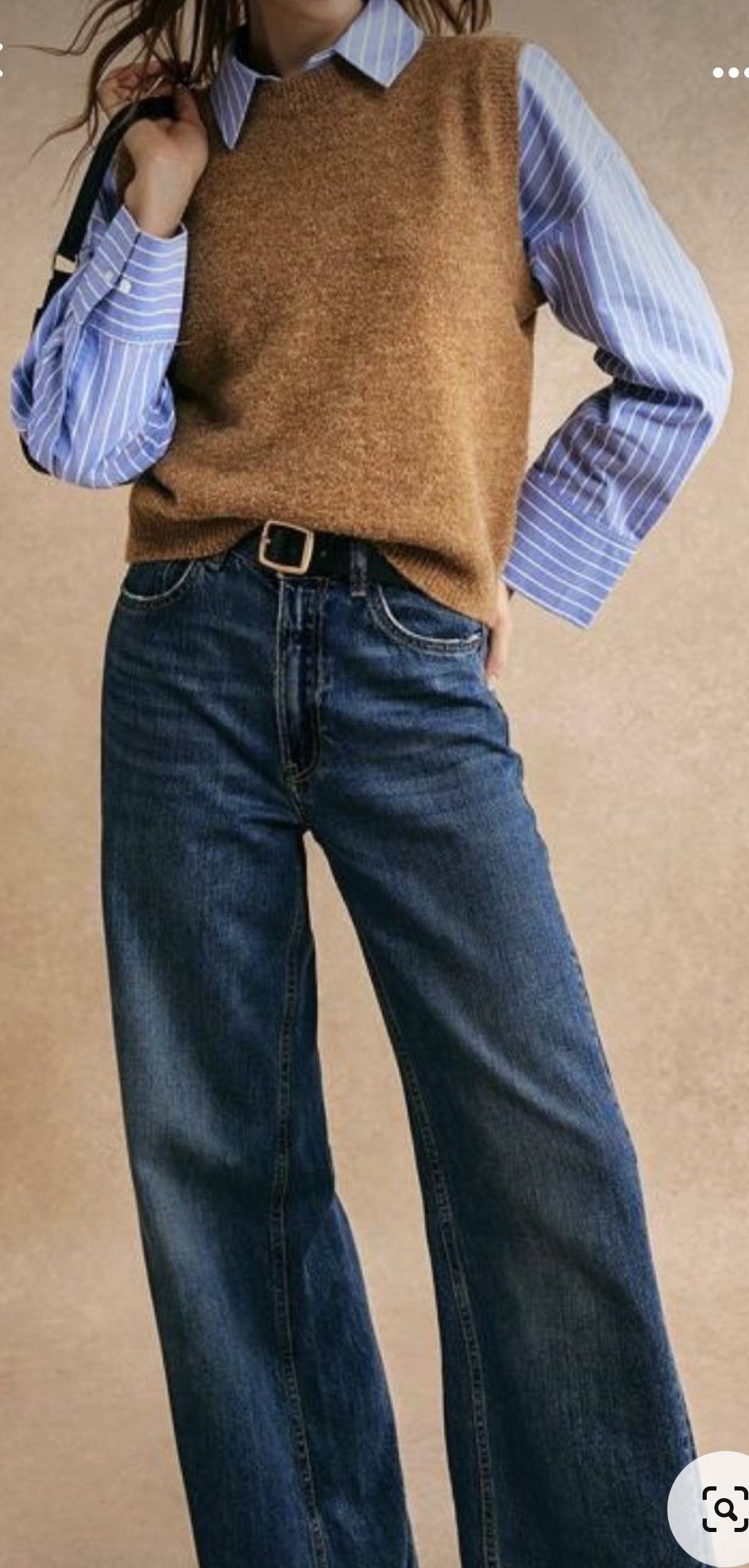 Person wearing a striped shirt under a brown sweater vest with blue flared jeans.