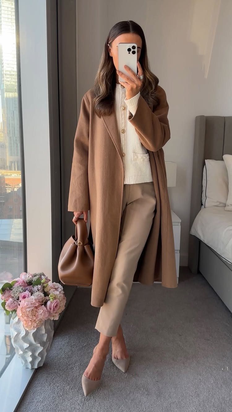 Woman in a tan coat, white sweater, and beige trousers posing for a mirror selfie.