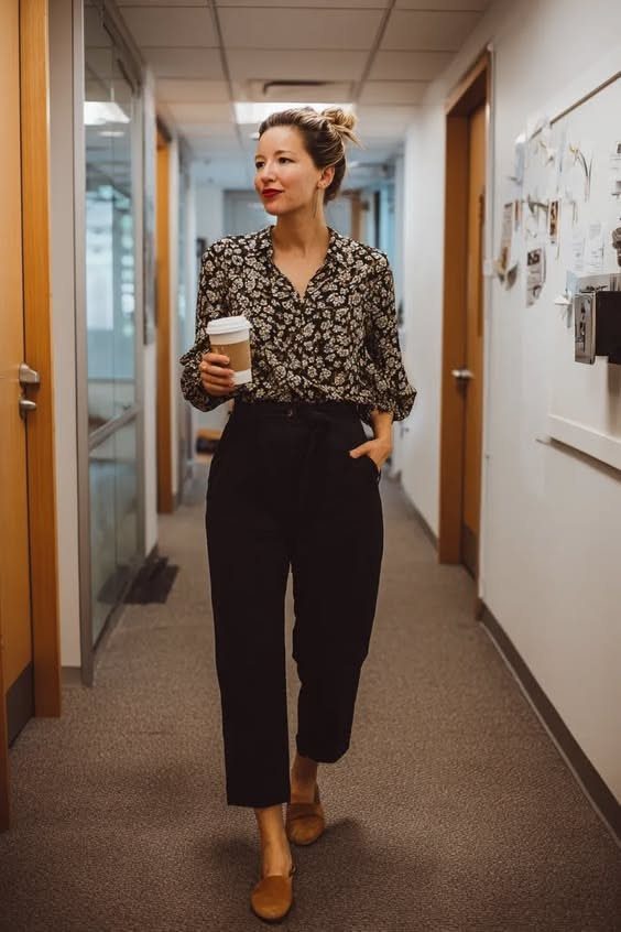Woman in patterned blouse and black trousers walking through an office hallway with a coffee cup.