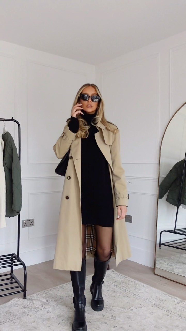Woman wearing a black dress, beige trench coat, and black boots standing in a white room.