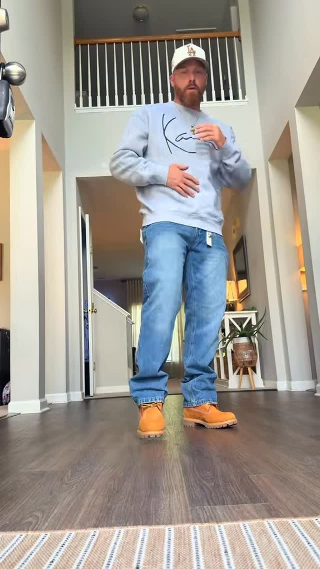 A man wearing a grey sweatshirt, blue jeans, and tan Timberland boots standing indoors.