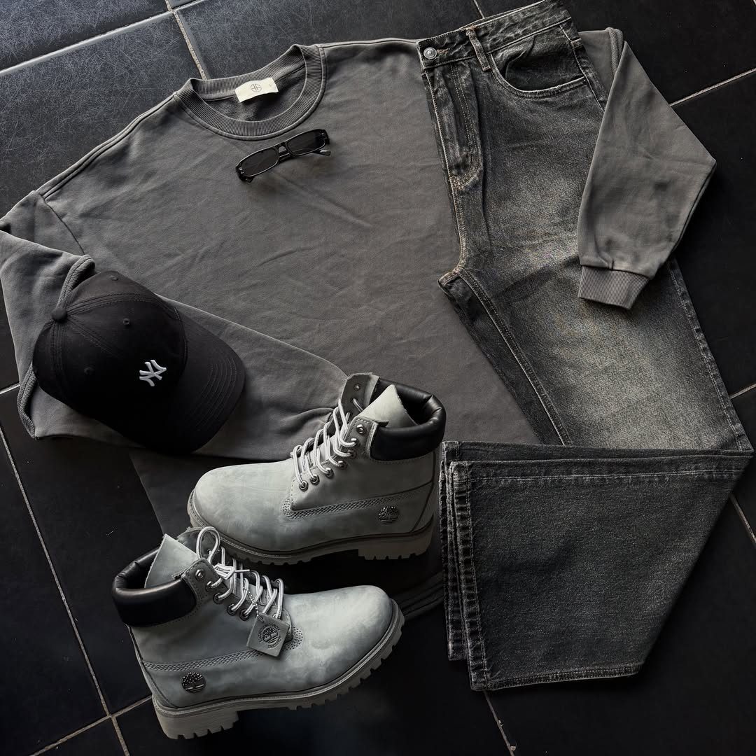 Grey long-sleeve shirt, denim jeans, black cap, sunglasses, and grey boots laid out on dark tiles.