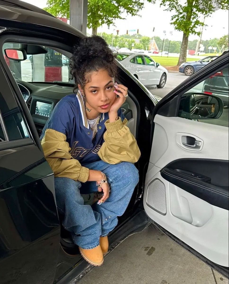 A young woman wearing a varsity jacket, blue jeans, and tan boots sitting in a car doorway.
