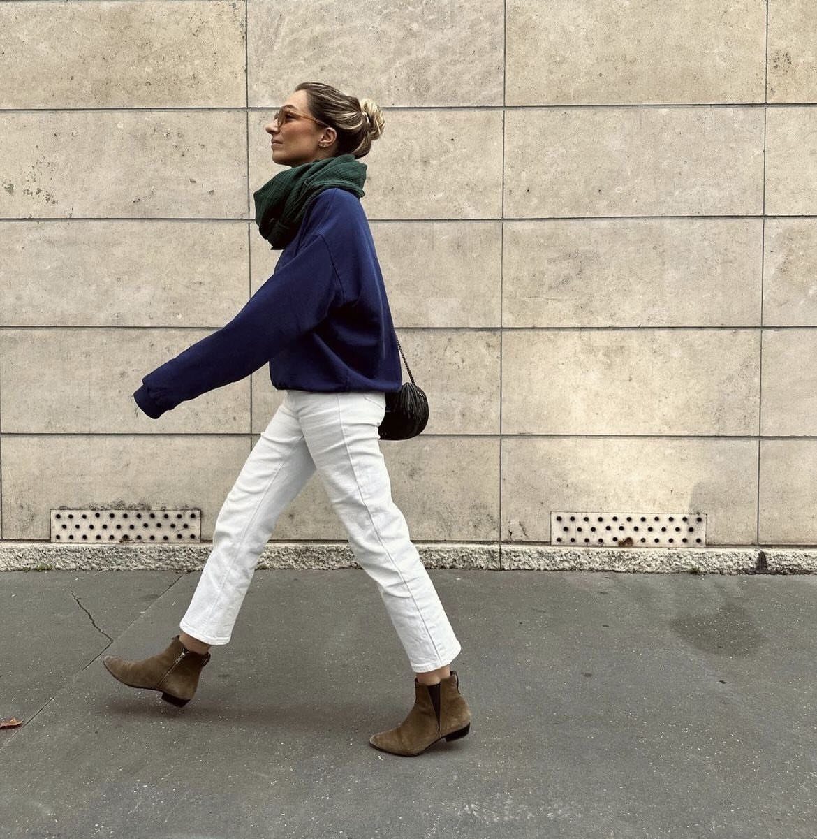 A woman in white jeans and brown suede ankle boots walking past a stone wall.