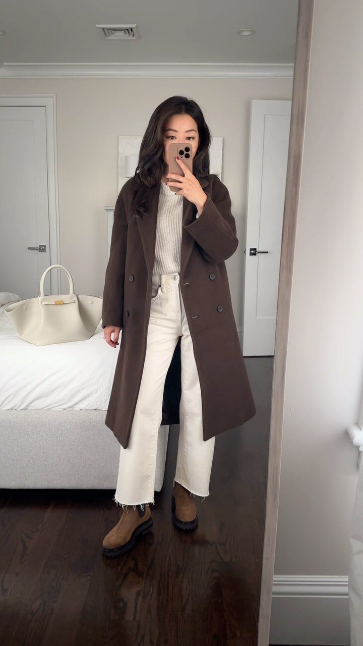 Woman in a brown coat, white sweater, white jeans, and brown boots posing in a mirror.
