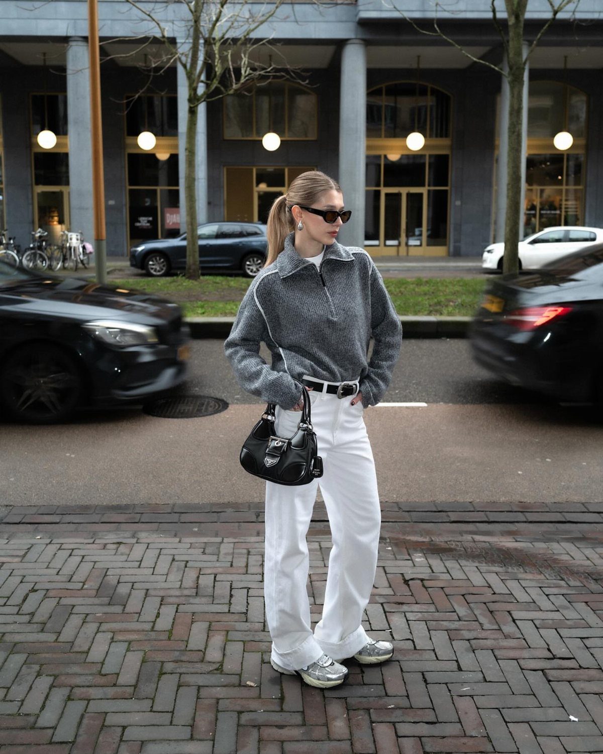 Woman wearing white wide-leg jeans and a grey chunky knit sweater standing on a city street.