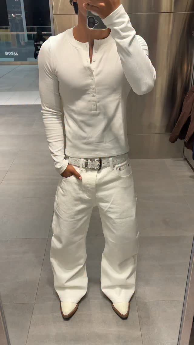 Man posing in a mirror wearing a monochrome all-white outfit with wide-leg jeans.