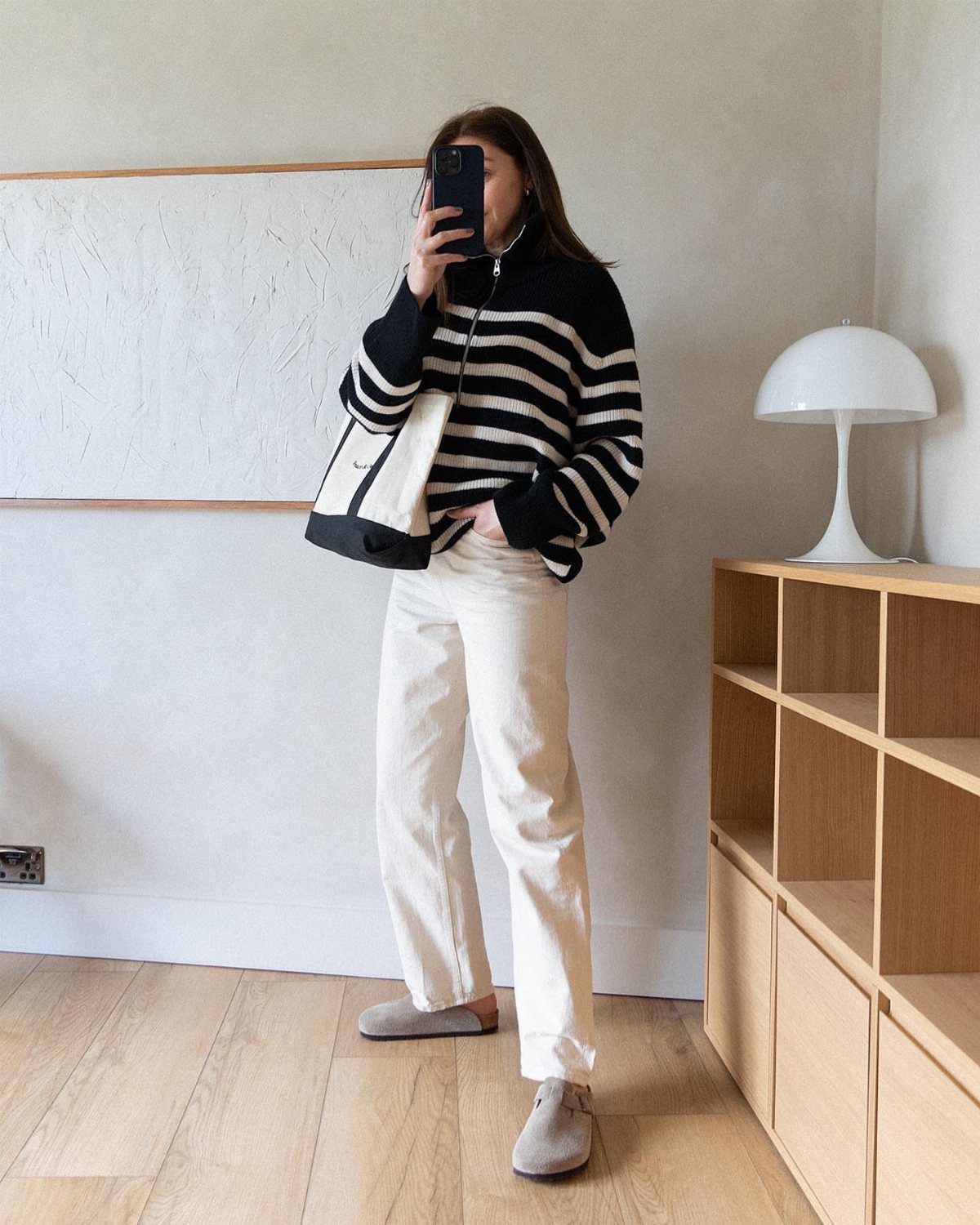 A person wearing a striped sweater and white jeans posing for a mirror selfie indoors.